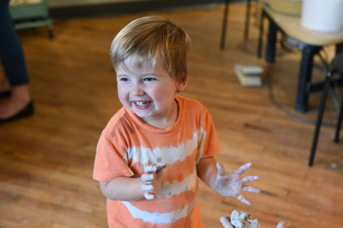 Smiling small child in an orange shirt with clay-covered hands.
