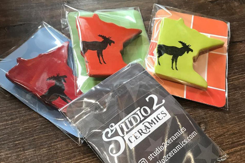 Four packages of state magnets. Three Minnesota shaped magnets (in red, orange and yellow with a stamped moose) and one is the back of the package with the Studio 2 Ceramics logo.