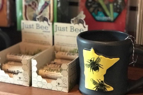 Cropped image of a dark gray ceramic mug decorated with a yellow Minnesota state cutout and bee stamps next to packages of "Just Bee" lip balm.